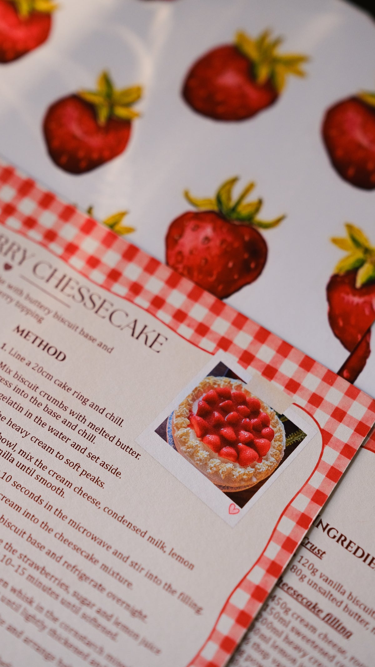 A strawberry cheesecake recipe card laid on red gingham fabric, beside a slice of cheesecake.